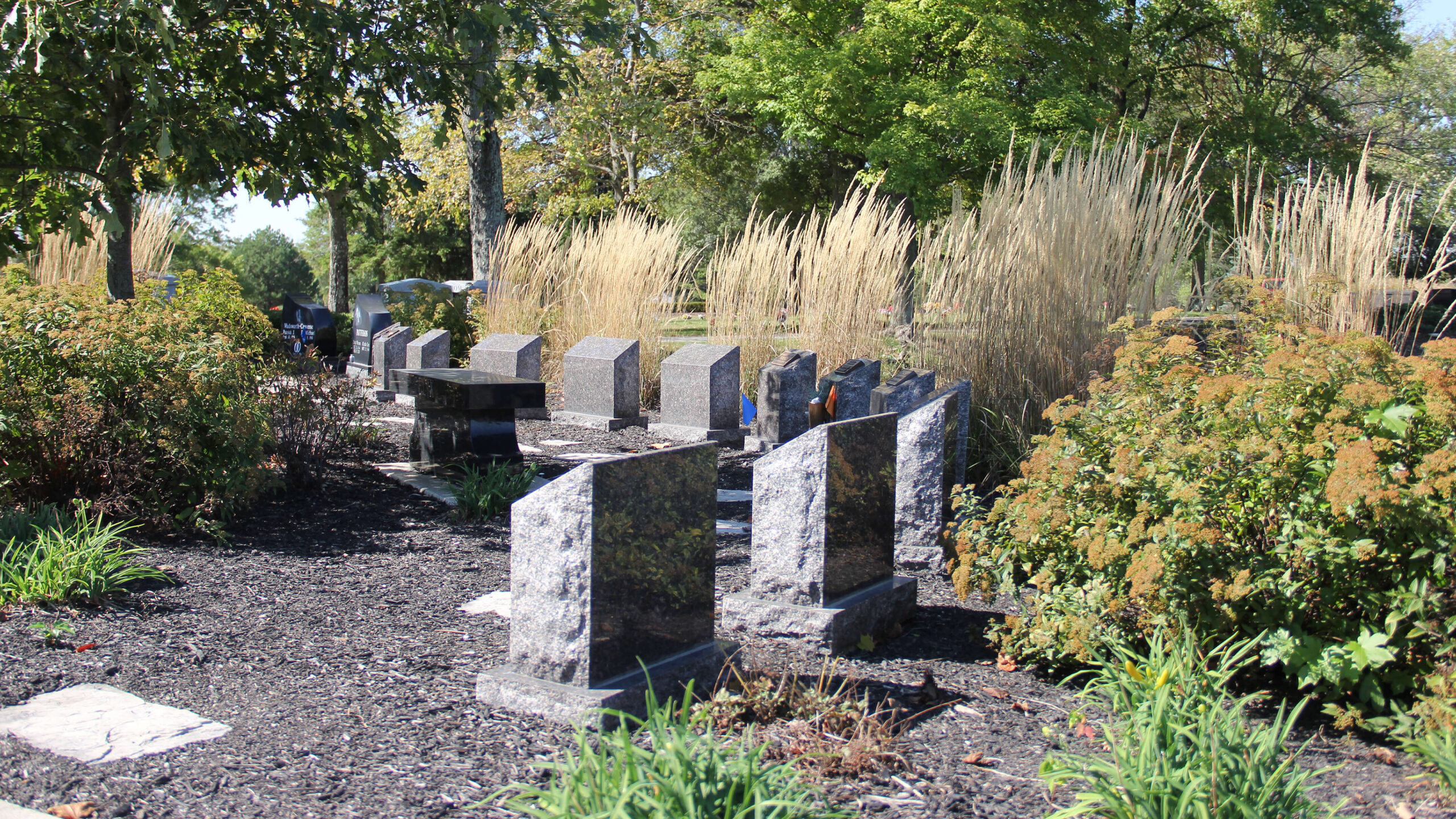 Cremation pillars arranged along a path in a cremation garden with bushes and trees providing shade and tall ornamental grasses along the pond in the background.