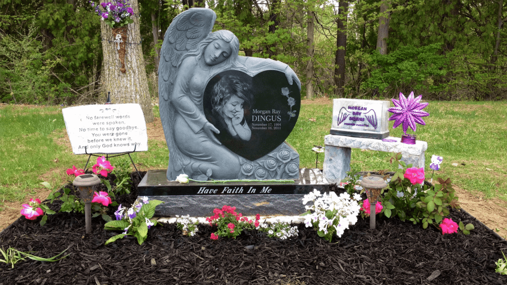Angel holding Jet Black heart with laser etched portrait of woman sitting in a plot surrounded by other signs and flowers.